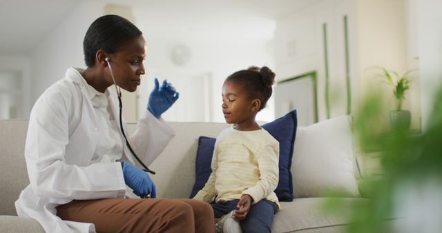 Doctor in white coat wearing blue gloves and using stethoscope to examine young girl in casual wear on living room couch. Perfect for illustrating healthcare services, pediatric home visits, doctor-patient interaction, and child health care.
