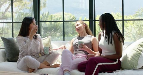 Three diverse women chatting and sipping coffee on sunlit sofa with cozy cushions