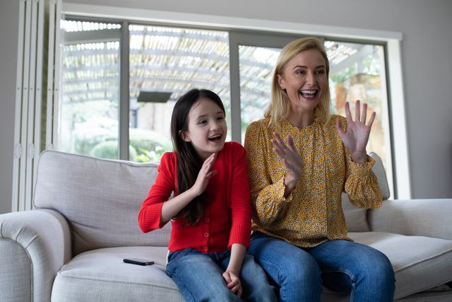 Caucasian mom and her daughter sitting on the couch at home. both of them are waving their hands. on the couch beside her daughter is a smartphone.