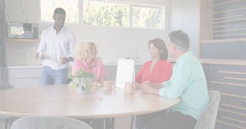 Serving man pouring drinks while friends chatting around round wooden table in kitchen