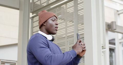African American commuter standing at shelter using smartphone with headphones and beanie