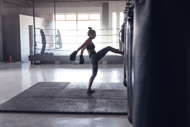 Side view of caucasian young female boxer wearing gloves kicking punching bag in health club. Sunlight, dedication, practicing, unaltered, boxing, sport, training, strength and fitness concept.
