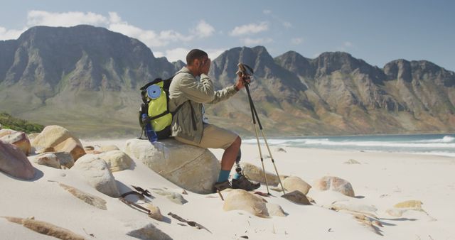 Biracial man with prosthetic leg trekking with backpack and walking poles taking a break on a beach. Long distance walking, fitness, challenge, disability, nature and healthy outdoor lifestyle.
