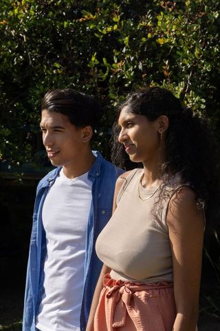 Young couple standing in sunlit garden looking left, casual summer fashion, gold jewelry