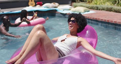 Young black woman relaxing on pink pool float laughing with friends backyard summer