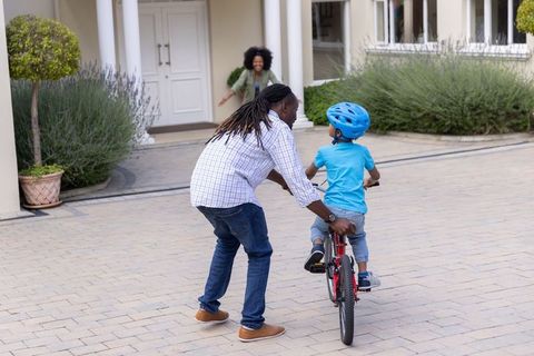 Father steadying child learning to ride red bike on driveway with mother cheering