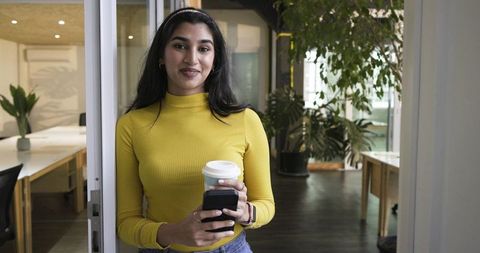Indian professional woman standing in coworking doorway holding coffee cup and smartphone