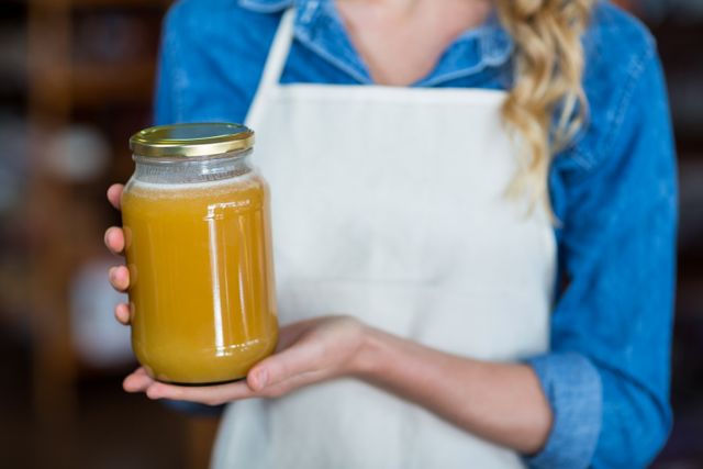 Female staff member in a supermarket holding a jar of honey. She is wearing a denim shirt and a white apron. This image can be used for promoting organic and natural food products, grocery stores, healthy lifestyle choices, and retail environments.