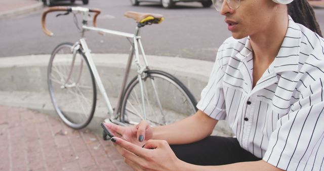 This image depicts a man sitting on a curb using a smartphone with a bicycle beside him in an urban area. He is dressed casually, possibly taking a break from cycling or waiting for someone. This provides a modern and relaxed setting, ideal for themes related to technology, communication, urban lifestyle, or fitness. It can be used for promoting mobile apps, cycling gear, or articles about urban living and tech usage.
