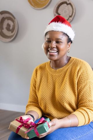 African american woman smiling and holding wrapped christmas gifts while wearing santa hat