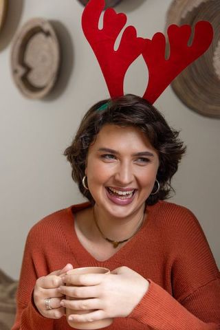 Young woman laughing holding mug wearing red reindeer antlers cozy holiday morning warmth