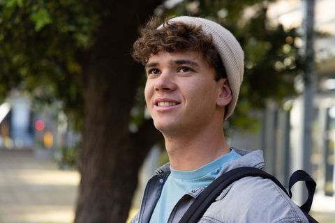 Urban student wearing beanie and backpack looking upward with hopeful smile on sidewalk