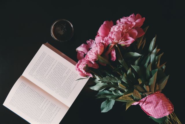 Bouquet of pink peonies lying next to an open book and a cup of coffee on a dark surface, creating a serene and peaceful atmosphere. Ideal for use in floral, literature, and lifestyle blogs, social media posts related to relaxation, and decorative or inspirational content.
