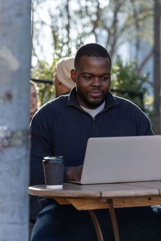 African american man working remotely on laptop at outdoor cafe table with coffee cup