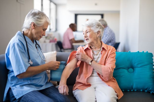Female doctor checking blood pressure of senior woman while sitting on sofa in nursing home. Ideal for use in healthcare, elderly care, medical services, and nursing home promotional materials. Highlights the importance of regular health monitoring and professional medical care for seniors.