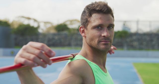 Male athlete wearing green shirt holding javelin on shoulders on sports field. Expression is determined. Ideal for advertising sports equipment, promoting athletic events, demonstrating javelin techniques, or depicting outdoor athletic training.