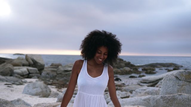 Young African American woman in a white dress dancing and smiling on a rocky beach at sunset. Suitable for promoting carefree lifestyles, summer getaways, vacations, and beachside activities. Perfect for advertisements, travel brochures, personal blogs, and social media content, emphasizing joy, natural beauty, and relaxation.