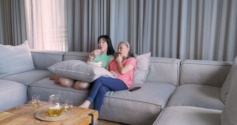 Asian Grandmother and Granddaughter Sharing Popcorn on Comfortable Sofa