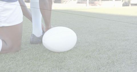 Kneeling rugby teammates adjusting cleat beside white ball on grass, teamwork moment