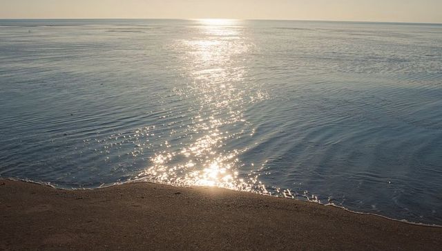 Sunlight reflecting on calm sea creating glitter trail toward sandy shore at sunrise