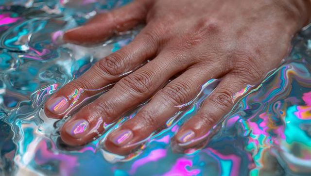 Pressing hand on iridescent liquid showing glossy manicure and neon rainbow ripples