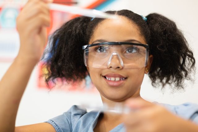 Close-up of smiling biracial elementary schoolgirl performing chemistry practical in lab. unaltered, education, laboratory, stem, scientific experiment, protection and school concept.