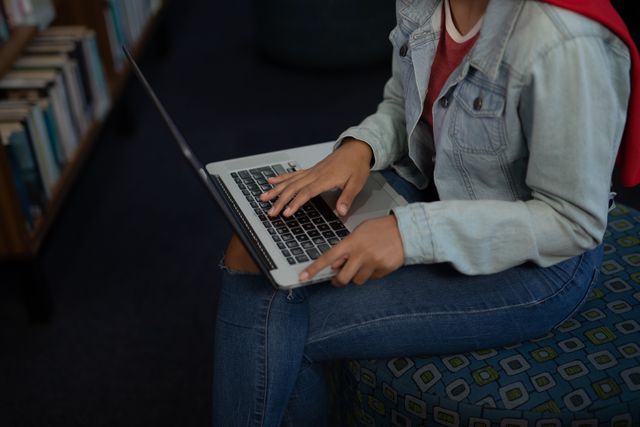 Asian female student in denim jacket and jeans typing on laptop while sitting between bookshelves in a library. Ideal for use in educational content, library promotions, academic resources, online learning platforms, and articles about student life.