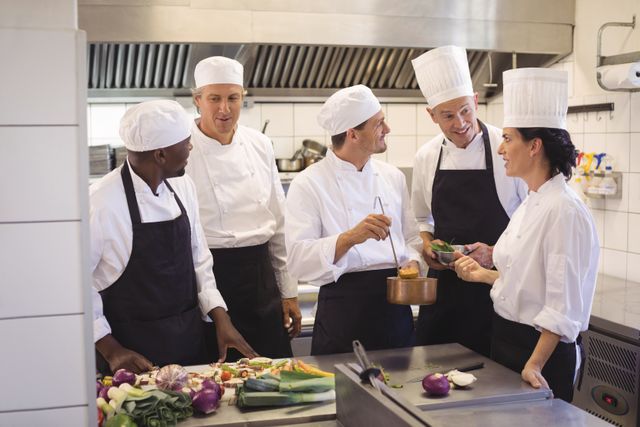 Group of chefs working together in a commercial kitchen, tasting and discussing food preparation. Ideal for use in articles or advertisements related to culinary arts, restaurant management, teamwork in professional kitchens, and the hospitality industry.