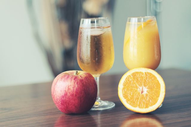 Refreshing drinks in tall glasses, one with apple juice, the other with orange juice. Ice cubes floating in the juices. Fresh apple and sliced orange displayed on table. Ideal for promoting healthy lifestyle, beverage choices, breakfast setups, or summer refreshment concepts.
