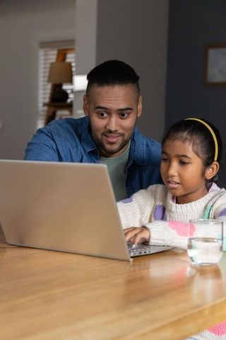 Diverse man guiding indian girl typing on laptop at wooden table during home learning