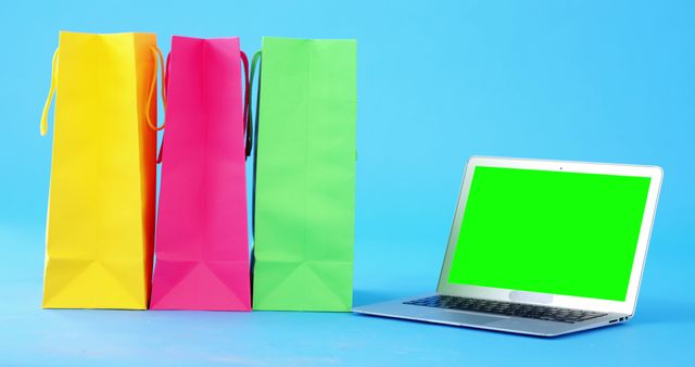 Three crisp, brightly colored shopping bags standing next to an open laptop with a green screen on a vibrant blue background. Ideal for illustrating themes of online shopping, e-commerce, retail technology, and digital purchases. Perfect for websites, advertisements, social media posts, and promotional materials highlighting shopping, retail, and tech-savvy marketing.