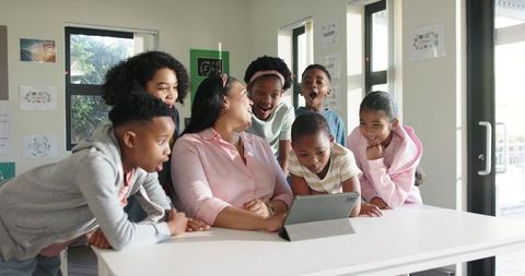 African American teacher leading elementary students around tablet in bright classroom