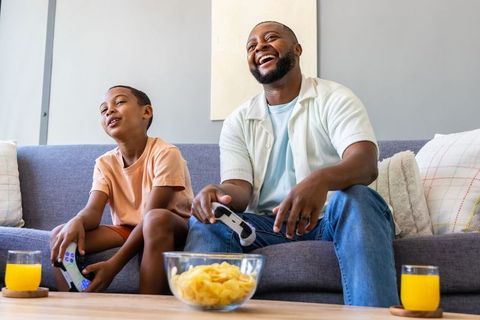 African american father and son playing video games together on couch with snacks