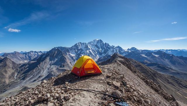 High-altitude camp on rugged alpine ridge featuring yellow-red tent and snow-capped peaks