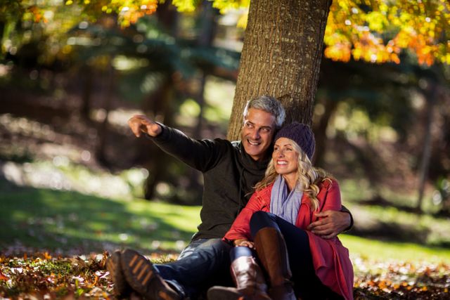 Middle-aged couple enjoying a peaceful moment under a tree in a park during autumn. The vibrant fall foliage and natural setting create a serene atmosphere. Ideal for use in advertisements, lifestyle blogs, and promotional materials focusing on relationships, outdoor activities, and seasonal themes.