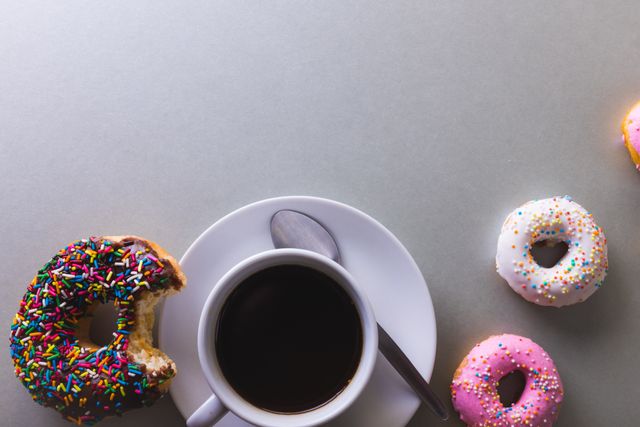 This image shows a cup of black coffee on a white plate with a spoon, surrounded by colorful donuts with sprinkles. The composition is from a top-down perspective, providing ample copy space on the white background. Ideal for use in food blogs, breakfast menus, coffee shop promotions, or advertisements for sweet treats and desserts.