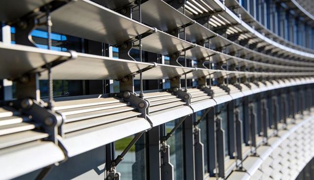 Curved metal louvers casting sharp shadows on glass facade with pivot brackets
