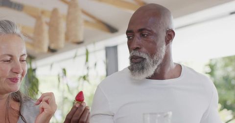 Senior african american man offering strawberry to gray-haired partner on sunny patio