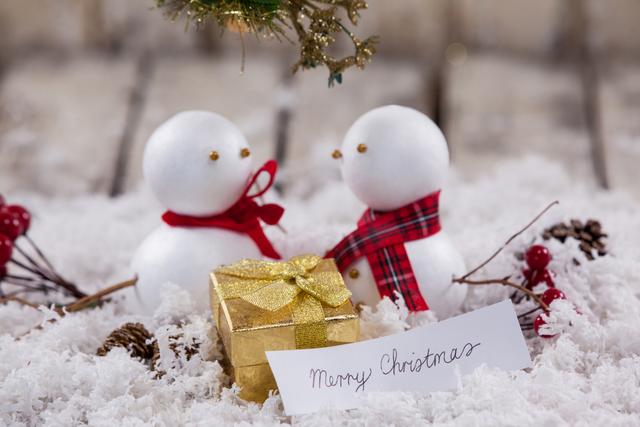 Two snowmen are standing in a snowy scene with a gold-wrapped gift and festive decorations. This image is perfect for holiday greeting cards, Christmas advertisements, winter-themed promotions, and festive social media posts.