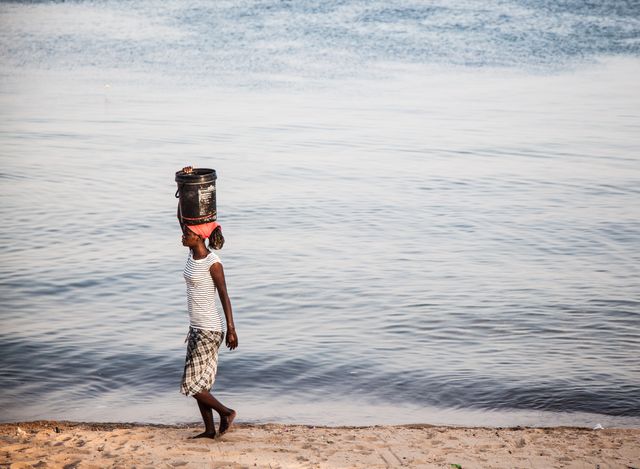 A child is seen carrying a water bucket on their head while walking along a riverbank at sunset. This scene represents traditional practices and daily life in rural African communities. It offers a glimpse into the cultural heritage and resilience of local populations. Ideal for use in educational materials, cultural and anthropological studies, and humanitarian campaigns.