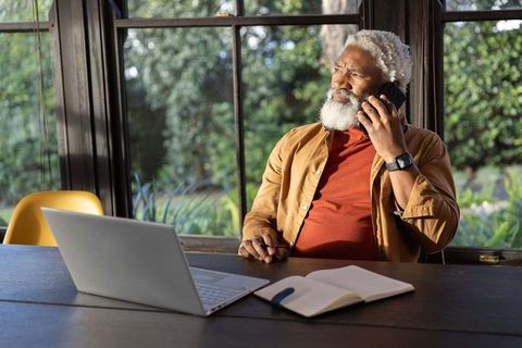 Senior african american man talking on phone at home workspace with laptop and notebook