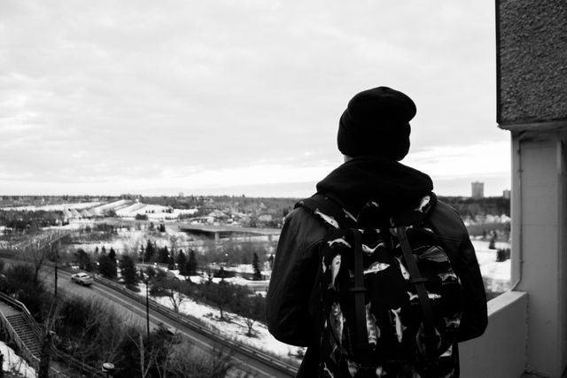 Person wearing dark winter clothing and a backpack is overlooking a snowy cityscape from an elevated position. Ideal for articles or advertisements about winter travel, urban exploration and adventure, outdoor activities in cold weather.