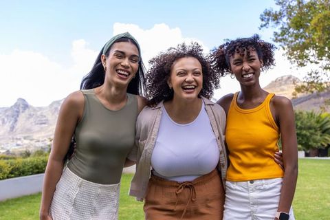 Three diverse women laughing and embracing outdoors on sunny lawn, friendship moment