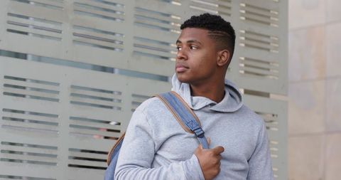Thoughtful african american man standing with backpack by modern urban facade in hoodie