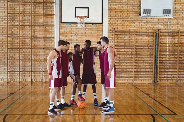 Basketball players in maroon uniforms huddling on an indoor court, discussing strategy and motivating each other before a game. Ideal for use in sports-related content, teamwork and motivation themes, athletic training materials, and promotional materials for basketball events.