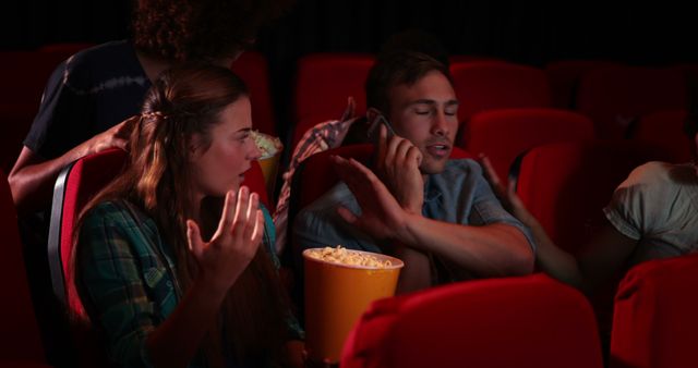 A diverse group of young adults is engrossed in a movie at a theater, with one person reaching into a popcorn bucket. Their expressions range from surprise to engagement, capturing the immersive experience of cinema.