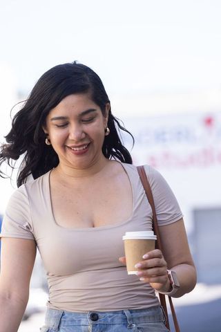 Smiling young woman walking with to-go coffee cup, wearing smartwatch and casual beige top