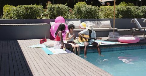 African american friends sitting poolside while man playing guitar at summer pool hangout