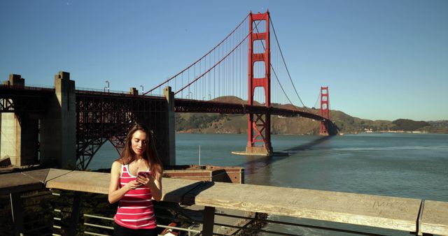 Woman in striped tank top using smartphone near Golden Gate Bridge in San Francisco. Suggests modern technology in travel and tourism. Ideal for promoting travel destinations, tech products, digital lifestyle.