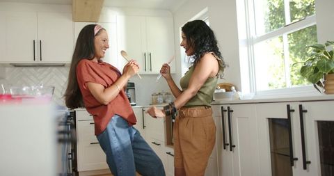 Indian and african american women singing and dancing in sunlit modern kitchen
